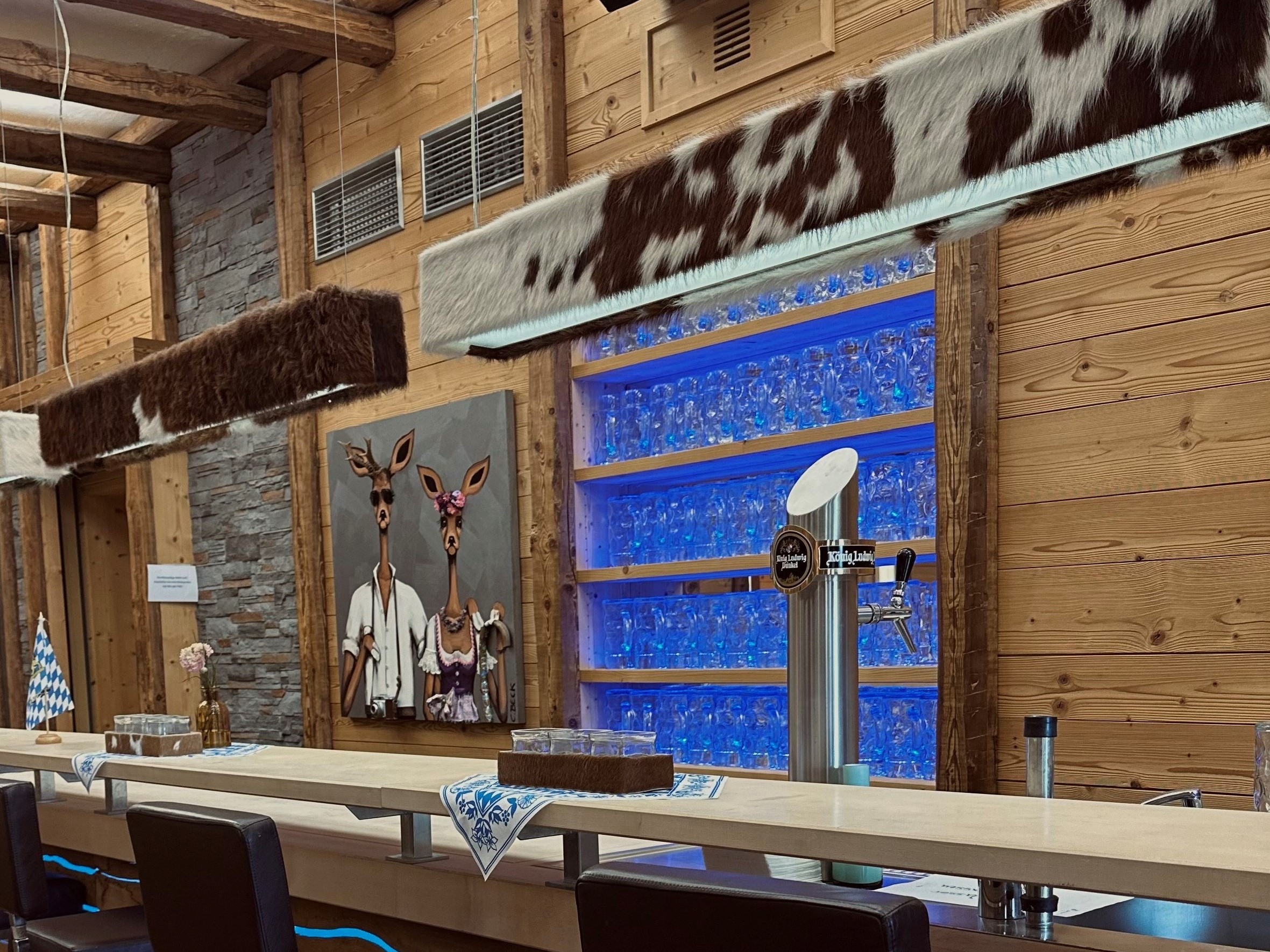 Der Indoor Biergarten: Holzbalken, mit Kuhfell bezogene Hängeleuchten, blau beleuchtete Regale voller Maßkrüge und eine Zapfanlage